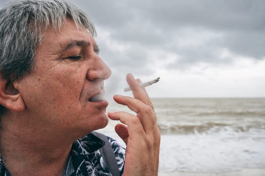 Lonely addicted sad elderly adult white man smokes a cigarette outdoors on the beach by the sea