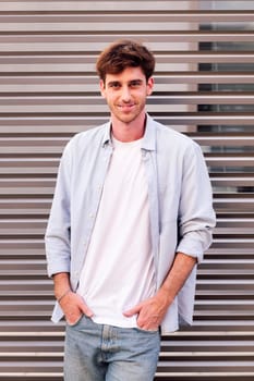 man in white shirt and blue jeans smiling looking at camera with hands in pockets, he is standing with a gray wall at background, concept of youth and urban lifestyle