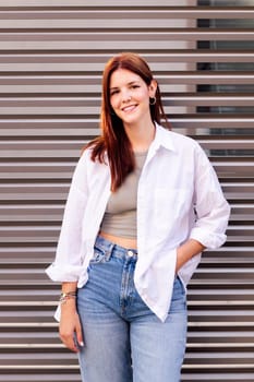 woman in white shirt and blue jeans smiling looking at camera with hand in pocket, she is standing with a gray wall at background, concept of youth and urban lifestyle