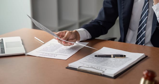 A lawyer is carefully examining contract documents while holding a pen, with a laptop placed on the desk in a modern office setting.