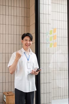 A young professional is joyfully celebrating success while holding a tablet, standing beside an organized array of colorful sticky notes on a glass wall in a contemporary office.