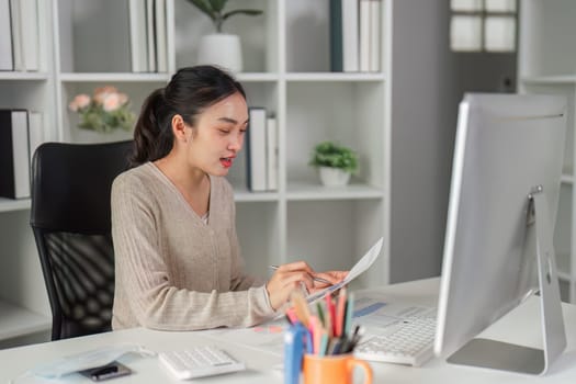 A young woman multitasks at her desk, reviewing reports and taking notes, showcasing efficiency in a professional environment.