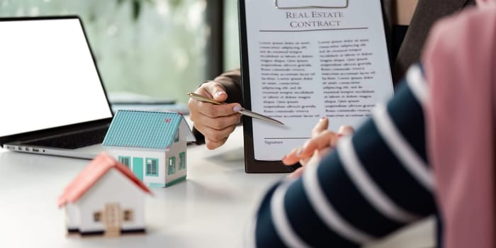 A professional real estate agent discusses a contract with a client, highlighting model houses and the agreement on the table.