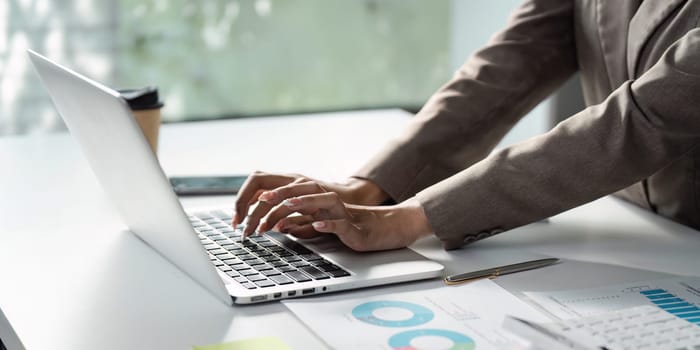A focused business professional of Asian descent types on a laptop, reviewing financial charts and reports, highlighting productivity and analysis in the workplace.
