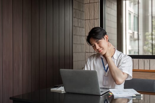 A young professional is rubbing his neck in discomfort while working on his laptop in a contemporary office, emphasizing the need for ergonomic practices.