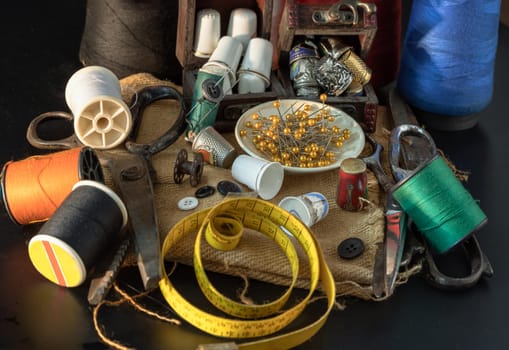 Spools of thread of various colors, Thimbles in small wooden treasure chest, Steel scissors, Sewing needles stuck in a green miniature spools thread and Measuring tape including pins in small round tray on Dark background. Sewing items set or  Sewing objects (Sewing notions), use it as your Wallpaper, Poster and Copy space, Selective focus.