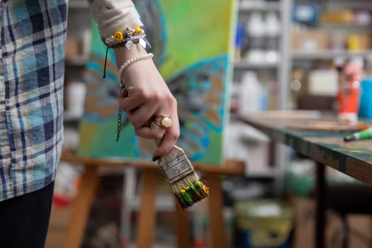 A closeup image captures a hand firmly grasping a paintbrush in an artists studio brimming with various supplies