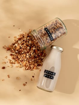 Granola and vegan milk. Healthy breakfast aesthetic concept. Homemade granola sprinkled of glass jar and vegan milk in glass bottle over champagne yellow or pastel beige background. Top view flat lay