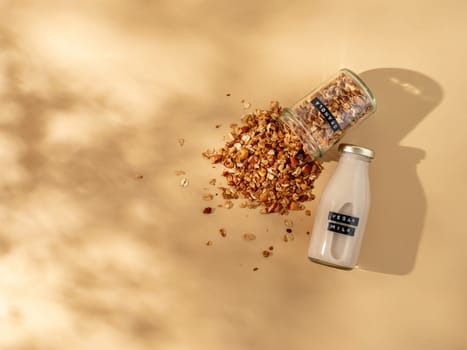 Granola and vegan milk. Healthy breakfast aesthetic concept. Homemade granola sprinkled of glass jar and vegan milk in glass bottle over champagne yellow or pastel beige background. Top view flat lay