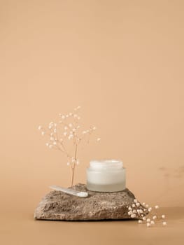 Cosmetic cream or moustirizer on stone over pastel beige background. Open round glass jar face cream and spatula. Copy space. Vertical. Mockup