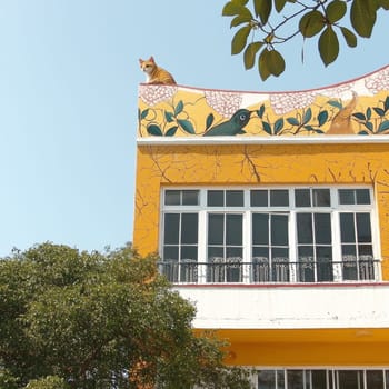 A ginger cat perches on the decorative roof of a yellow building. Ornate paintings of plants and birds adorn the building, framed by a sunny sky and verdant trees below.