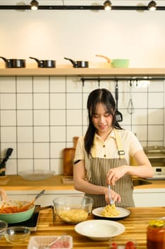 Young woman wearing apron twirls freshly cooked spaghetti onto a plate in a home kitchen.