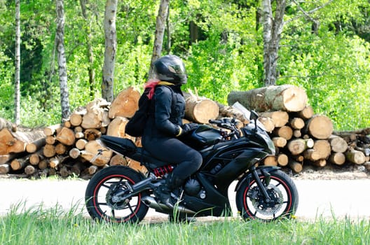 Biker girl in a black jacket on a motorcycle. Motorcyclist in outfit with motorcycle in sunglasses and jeans in summer. Side view. The girl has pink hair. With tree logs in the background