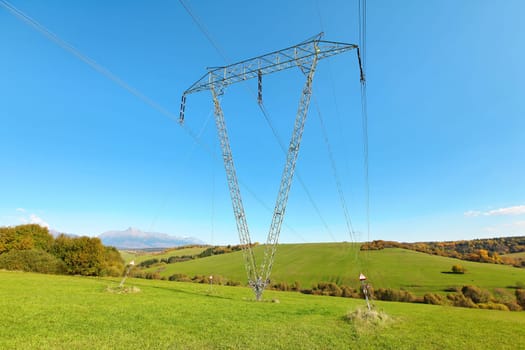 Large metal power pylon under electricity lines built in countryside with green grass covered hills and small forests, mount Krivan (Slovak symbol) at distance, blue sky background 