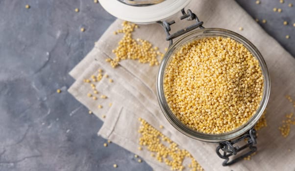 Glass jar of raw uncooked yellow millet grain on napkin on gray table top view, copy space. Ancient pseudocereal seeds. Organic super food. Healthy food concept. Gluten free alternative seeds