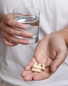 Woman in white t-shirt holding beige capsules in cupped hand and water glass in hand closeup. Female or male taking dietary supplements or vitamins. Healthcare concept