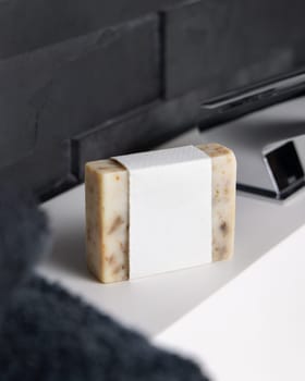 Beige handmade soap bar with blank label on washbasin near faucet and black bath towel in modern dark grey bathroom close up, mockup. Natural herbal product for daily male hygiene