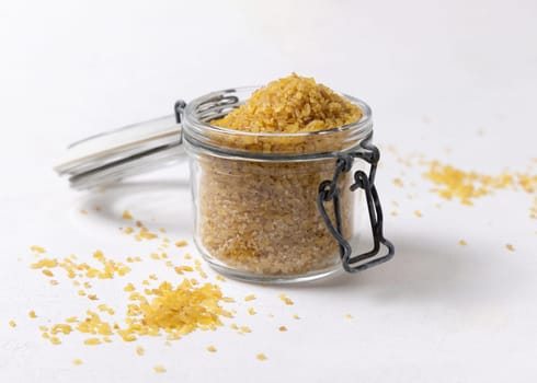 Opened glass jar of raw dry uncooked yellow bulgur grain on white table closeup, copy space. Middle Eastern cousine. Organic super food for healthy diet. Ancient alternative cereal. seeds