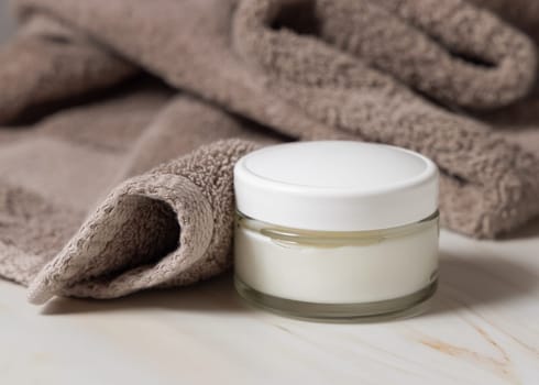 Cream jar with white lid near light brown bath towel closeup. Natural organic cosmetics for facial care. Lifestile scene of skincare, essence of Spa and Wellness, macro cream texture