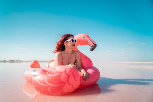 A woman wearing a swimsuit and sunglasses lays on a pink flamingo float in a pink lake. The sky is blue.