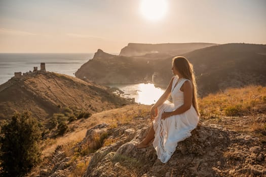 A woman in a white dress sits on a rock overlooking a body of water. The scene is serene and peaceful, with the woman enjoying the view and the calmness of the surroundings