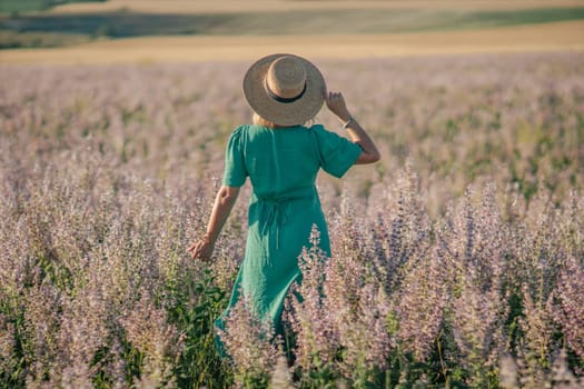 Here's a caption under 150 characters describing the image: Woman in teal dress and straw hat walks through a field of purple flowers. Golden hour light. Back view.
