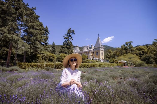 A woman is sitting in a field of purple flowers. She is wearing a white dress and a straw hat. The field is surrounded by trees and the sky is clear
