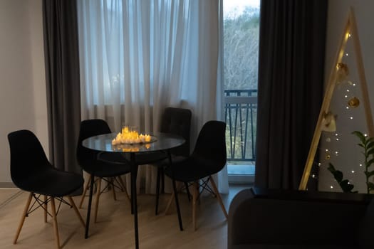 Cozy apartment interior: Round glass dining table with four black chairs, lit candles, dark curtains framing a balcony view, and a gold decorative ladder.