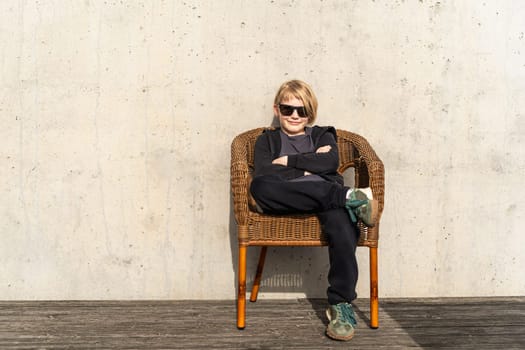 young boy sitting on wicker chair with crossed arms and legs against concrete wall.