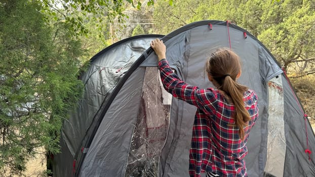 A young woman assembling a tent in an outdoor setting. She stretches the fabric and secures the structure, preparing for camping. Natural surroundings in the background.