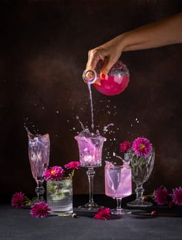 Hand pouring a vibrant pink beverage from a glass bottle into elegant crystal glasses, causing a splash and surrounded by ice cubes and purple flowers