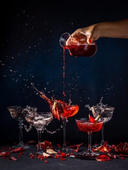 Red cocktail being poured into glasses, creating splashes and adorned with pomegranate seeds, on a dark background