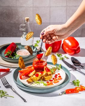 Chef pours spicy sauce from a spoon onto a plate of roasted potato wedges, red bell peppers, and chili peppers