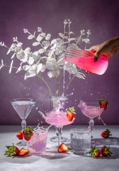 Pink cocktail being poured into glasses, creating splashes, decorated with strawberries and ice, with white flowers and a purple background