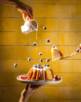 Chefs are pouring icing and serving a slice of delicious blueberry bundt cake decorated with grapefruit against a yellow tiled wall