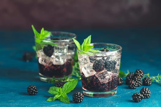 Two glasses of sparkling blackberry cocktail with ice cubes, fresh mint and blackberries on blue background