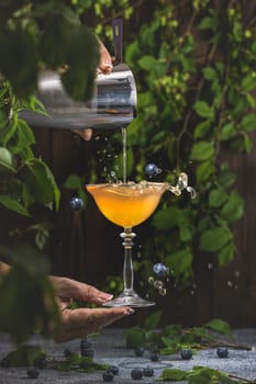 Bartender pours a vibrant cocktail into a stemmed glass, creating a splash with fresh blueberries amidst a lush, leafy backdrop
