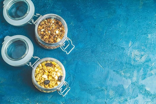 Two open glass jars containing chocolate and classic homemade granola on rustic blue background with copy space