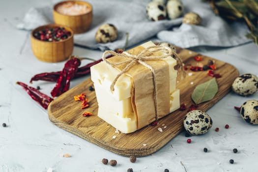 Maasdam cheese slices wrapped in brown paper and twine, arranged on a wooden board with quail eggs, dried chili peppers, and spices
