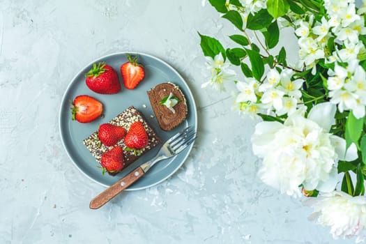Chocolate roll cake decorated with fresh strawberries, served on a gray plate with a fork, near a bouquet of white flowers