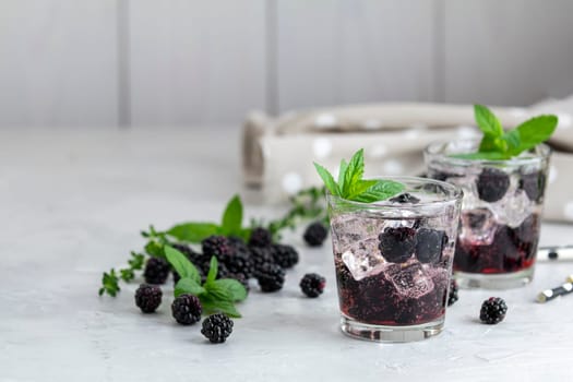 Two glasses of refreshing blackberry cocktail with ice, mint, and thyme are garnished on a bright kitchen table