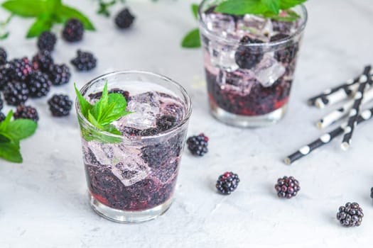Two glasses of refreshing blackberry cocktail with ice, mint and black and white straws on a light background