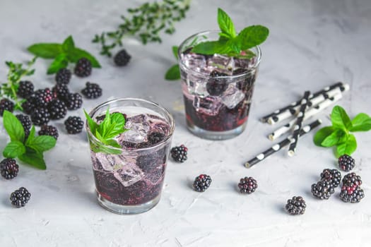 Two glasses of blackberry cocktail, garnished with mint and ice, sit among fresh blackberries, mint sprigs, and thyme on a textured surface