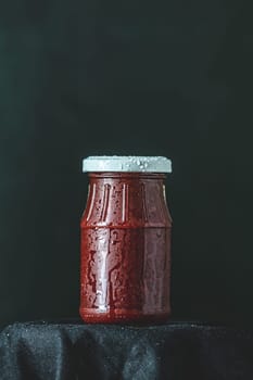 Small glass jar containing red sauce, covered with condensation, sits on a dark cloth against a black background
