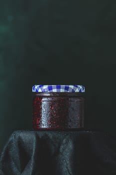 Glass jar of red raspberry jam with condensation and checkered lid sits on black cloth against a dark background