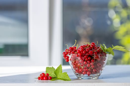 Ripe red currants spilling out of a glass bowl, accompanied by vibrant green leaves, create a refreshing scene on a white table