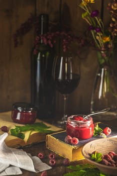 Homemade raspberry jam in a glass jar with fresh raspberries, mint leaves, a bottle of wine, a glass of wine, old books, and flowers on a wooden table