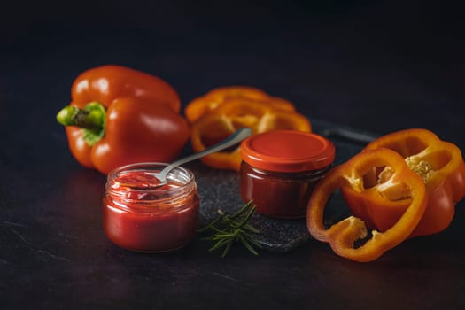 Homemade bell pepper sauce in glass jars with fresh, sliced bell peppers and rosemary on a dark background