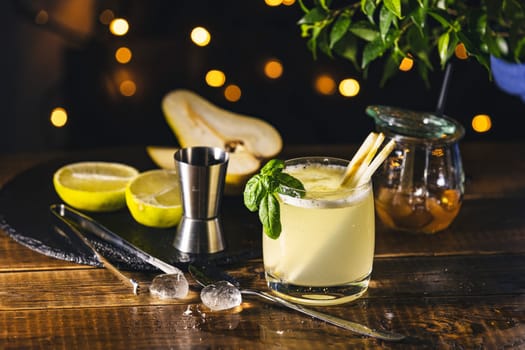 Refreshing cocktail being prepared with pear, lime, honey, and basil, featuring a jigger and ice on a wooden table with festive lights in the background
