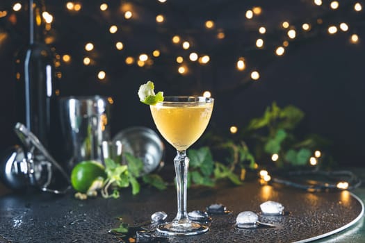Refreshing cocktail served in a coupe glass, garnished with a lime wedge and mint leaves, sits on a wet bar top amidst bar equipment and festive lights